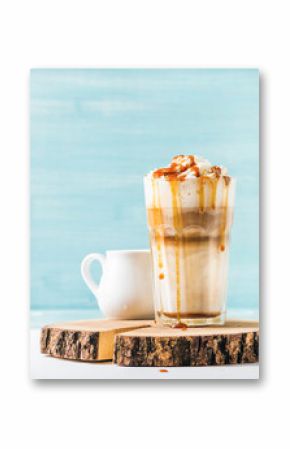 Latte macchiato with whipped cream and caramel sauce in tall glass on wooden board over blue painted wall background, selective focus, copy space, vertical composition