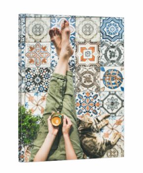 Lazy morning on terrace. Flat-lay of womans legs in cosy linen pants, plant, cat and cup of coffee in hand over colorful moroccan tile floor, top view