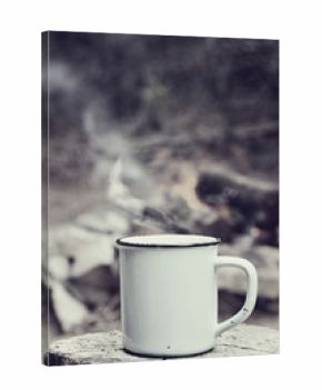 Cup of hot steaming coffee sitting on an old log by an outdoor campfire with a vintage antique edit. Extreme shallow depth of field with selective focus on mug.