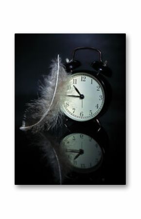 Black alarm clock on a mirror surface, feather on black background