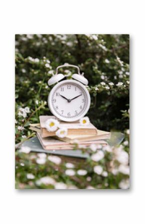 A retro alarm clock stands on a stack of books amongst the flowering branches of a shrub. A time to read and rest. A series of photographs about time and clocks.