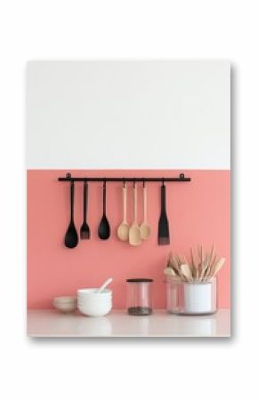Minimalist kitchen interior with pink walls, red hanging decor, and wooden teaspoons on black metal hanger. Transparent cutlery holder with knives and forks, soft peach background with plates and bowl
