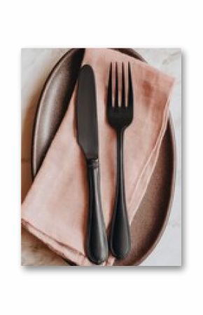 Flatlay of matte black knife and fork atop a blush pink linen napkin on a dark brown oval plate, set on a marble surface