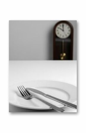 Empty white plate with utensils set on a white surface, in front of a blurred antique clock.