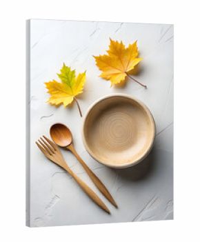 Overhead view of a rustic wooden plate and cutlery set alongside vibrant yellow autumn maple leaves on a textured white background