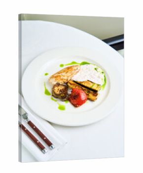 Grilled chicken is plated alongside roasted vegetables including zucchini, tomato, and a creamy herb sauce. Elegant dining setting features a white tablecloth and polished cutlery