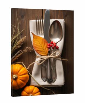 Autumnal Thanksgiving table setting: cutlery tied with twine on linen napkin, orange pumpkins, fall leaf, berries, wheat on dark wood background, symbolizing harvest, gratitude, and family gathering