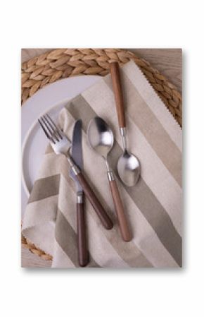 A modern cutlery set with wooden handles is elegantly arranged on a striped linen napkin atop a wicker placemat. The set includes a fork, knife, and two spoons, highlighting contemporary dining style.