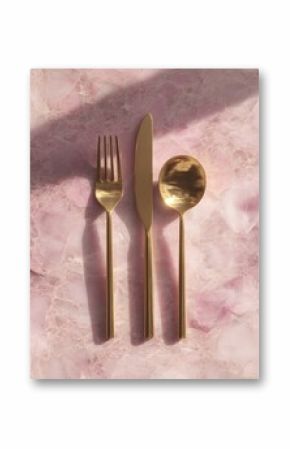 Gold cutlery set ? fork, knife, spoon ? arranged on a pink marble surface, sunlit