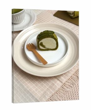A slice of matcha roll cake served on a white ceramic plate with green tea and wooden cutlery on a beige cloth.Bright,clean tabletop food photography ideal for dessert menus and bakery promotions.
