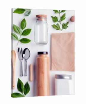 A collection of reusable items like a bamboo water bottle, glass jar, and cutlery are artfully arranged with fresh green leaves and a paper bag on a white surfa