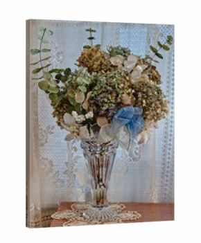 This vertical still life shows dried hydrangea and other florals in a crystal vase, on top of crochet doilies on a table, with old fashioned vintage lace curtains in a window behind.