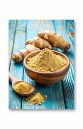 Ginger powder in a shallow bowl sits atop a slice of fresh ginger root on a blue wooden background
