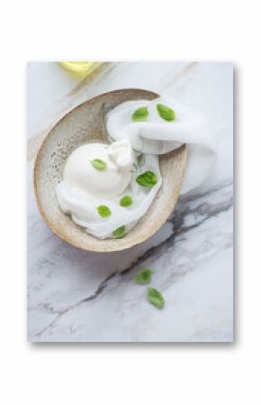 Bowl with burrata cheese in brine and green basil leaves, vertical shot on a white stone background, above view