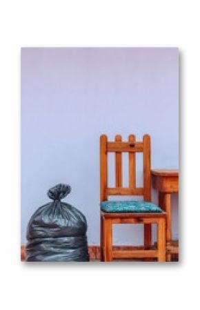 Stark Still Life: Dark Trash Bag Near Wooden Chair and Table Against Light Purple Wall