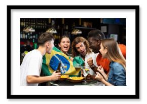 Multiracial Brazil sports fans, men and women, supporting their favourite team in bar, raising state flag and screaming chants together.