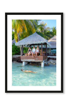 Three women relax on a wooden terrace above turquoise water while another swims nearby, tropical resort in maldives, peaceful vacation, summer retreat, relaxation