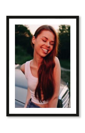 Young woman with a joyful expression in a casual outfit poses outdoors near a car during sunset, highlighting a carefree summer vibe.