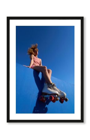 Young woman wearing roller skates sitting on blue wall under vivid blue sky