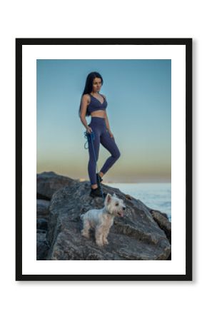 beautiful brunette woman wearing fitness attire stands on a rock by the beach She enjoys a serene sunset stroll with her white dog beside her, showcasing her smooth skin after epilation