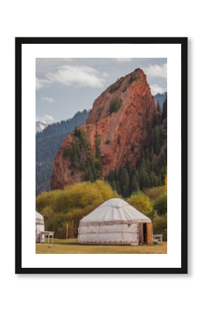 Traditional yurts stand in a picturesque valley, framed by towering red rock formations and lush evergreens. The stunning autumnal foliage adds a vibrant touch to this breathtaking scene in