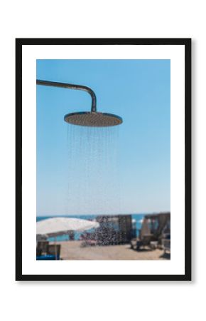 A metal shower head dispenses water in a beach setting with a bright blue sky above. Sunbeds and umbrellas are visible near the shore, inviting relaxation and refreshment