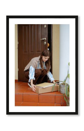 A young woman wearing a sweater and striped shirt bends down to pick up a package from her front porch. The setting is a warm, inviting home with potted plants.