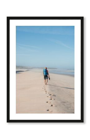Lonely barefoot backpacker man walking by sandy Portugese beach in morning on the Atlantic ocean coast. Shot during the Camino Portuguese - popular pilgrimage route. Active people, traveling concept.
