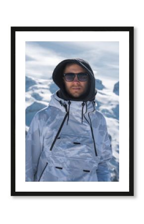 A man in a white and gray camouflage ski jacket with a hood and sunglasses stands in a snowy alpine environment with peaks and a partly cloudy sky.