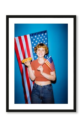 A young person smiles brightly while holding a hot dog and an American flag. The vibrant blue background and festive atmosphere create a cheerful vibe, capturing the essence of summer celebrations.