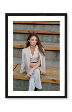 stylish woman in light suit sitting on wooden bleachers outdoors with calm expression, casual fashion, spring season, natural light, modern casual style