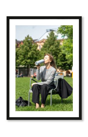 Exhausted woman squinting from heat, drinking water thirstily resting on grass in park after work. Refreshment, female on chair with beverage, park relief, hydration and recovery, sweltering weather