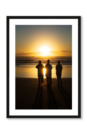 Three teenage girls silhouetted quietly on the dark sands of Muriwai Beach, gazing at a golden sunset as it reflects on the ocean waves and clouds in a serene evening atmosphere