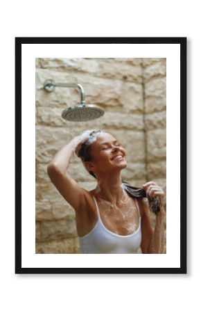 smiling woman washing hair under outdoor shower with stone wall background, enjoying warm water and natural light, fresh and relaxed morning moment