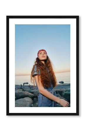 young woman with long curly hair wearing a red bandana and denim outfit enjoying sunset at rocky beach with calm sea and clear sky in warm evening light