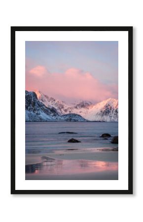 Colorful winter sunrise at Haukland Beach, Lofoten Islands, Norway. Snowy mountain and pink clouds reflect in the calm sea and wet sand, creating a peaceful Arctic landscape.