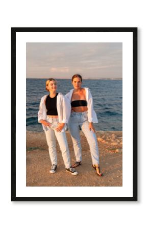 Women Beach Sunset Two women pose on a rocky beach at sunset, wearing white shirts and jeans.