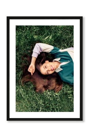 Positive woman with long brunette hair laying on green grass outdoors wearing casual stylish clothes during fall