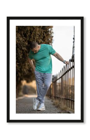 middle aged european man dressed in a turquoise t shirt and pink sweatpants walks thoughtfully along a park path next to a decorative fence surrounded by greenery during daytime.