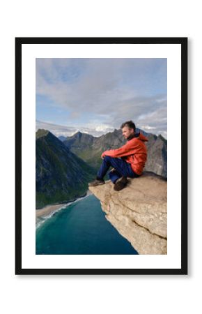 A hiker sits on the edge of a rocky precipice on the Ryten Kvalvika Beach Trail, admiring breathtaking views of the turquoise waters and rugged landscape of Lofoten, Norway.