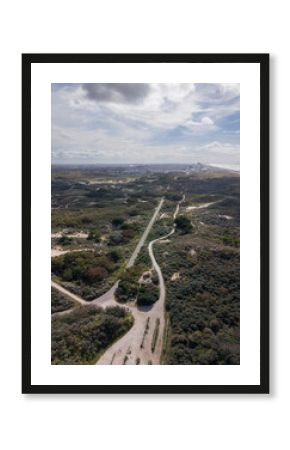 Aerial view of a coastal zone with winding paths through sandy dunes and dense vegetation, leading toward a shoreline cityscape beside the ocean under a partly cloudy sky.