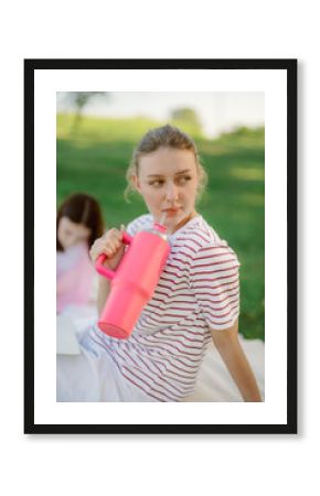 A young woman leans back while sipping a bright pink drink from a large cup, enjoying a picnic on a sunny day in a park with green grass. Another person is seated nearby.