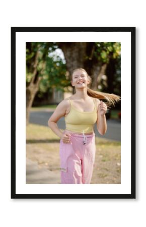 A young woman with long hair jogs with a smile along a tree-lined street. She wears a light yellow top and pink pants, enjoying a warm day outdoors.