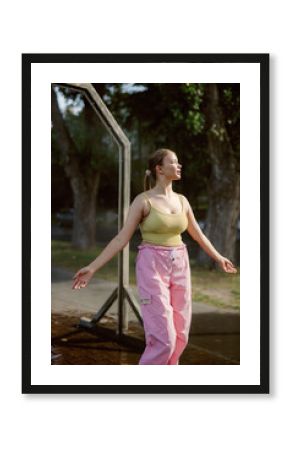 A woman stands in a park, soaking up the sun while wearing a yellow top and pink pants. She smiles and spreads her arms, enjoying a joyful moment in nature on a warm day.