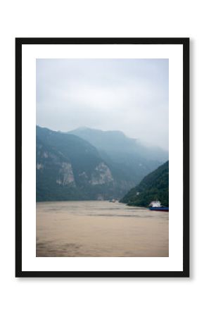 Yichang Three Gorges in China features a wide river flowing between immense, green mountains under a cloudy sky with boats navigating the water.
