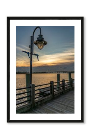 A peaceful sunset illuminates the Wilmington, North Carolina Riverwalk, where a vintage lamppost stands over a wooden boardwalk beside the calm Cape Fear River.