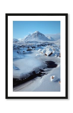 Bright, freezing winter scene in Glencoe featuring the iconic Buachaille Etive Mor rising above the River Etive. The frozen river and snow-covered rocks dominate the foreground under a clear blue sky