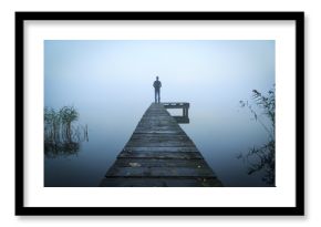 Man standing on a jetty at a lake during a foggy, gray morning.