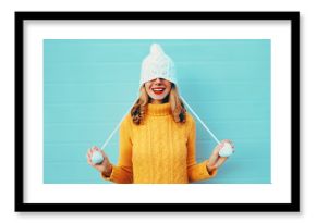 Portret zimowy szczęśliwy uśmiechnięta młoda kobieta zabawy ciągnie kapelusz na oczy, ubrany w żółty sweter z dzianiny i biały kapelusz z pom pom na niebieskim tle ściany