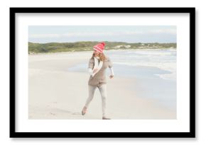 woman running on white sandy beach wearing red knit cap, white scarf, with green dune backdrop